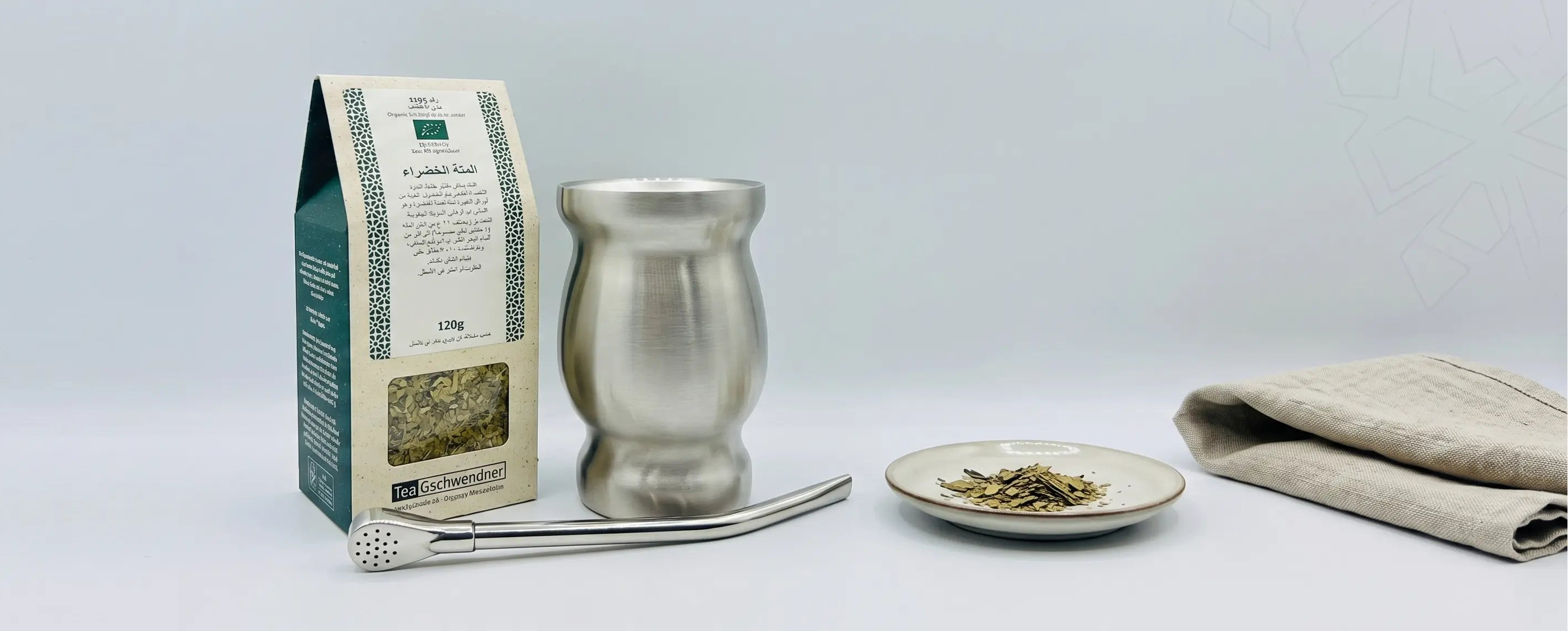 A silver metal tamper and matching metal cup set displayed next to a paper packaging box, a small plate with loose tea leaves, and a folded beige cloth on a light gray background.