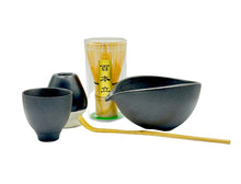 Tea-making set with black ceramic bowls, a glass container, and wooden sticks on a white background