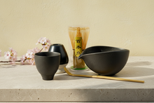 Tea-making set with black ceramic bowls, a glass container, and wooden sticks on a white background