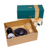 A tea gift set displayed in an open tan box on a dark table, with the matching teal and gold box behind it. The set includes a bamboo whisk, a dark ceramic teapot or scoop, a small bottle with a white label, and a small wooden utensil.