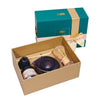 A Japanese matcha tea set gift set displayed in an open tan box, with its teal and gold branded gift box standing behind it on a dark table against a patterned wallpaper background.