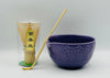 Purple ceramic bowl with a wooden spoon and a transparent container with matcha sticks on a gray background